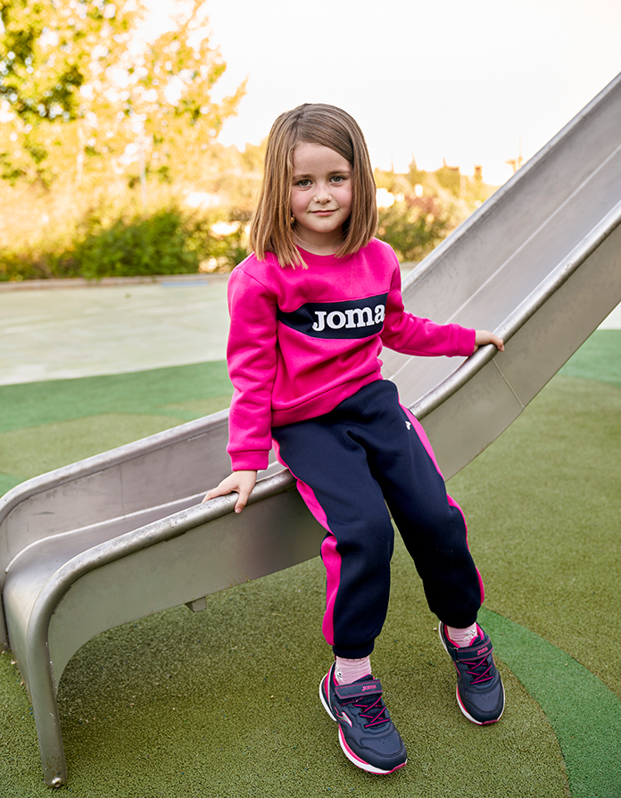 Fille vêtue d’un survêtement scolaire Stripe de Joma assise sur un toboggan.